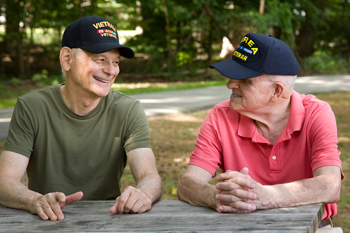Two elderly veterans wearing military caps sit together at an outdoor picnic table, smiling and talking with each other