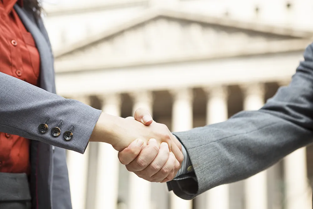 Two professionally dressed individuals shake hands in front of a government or civic building with large columns